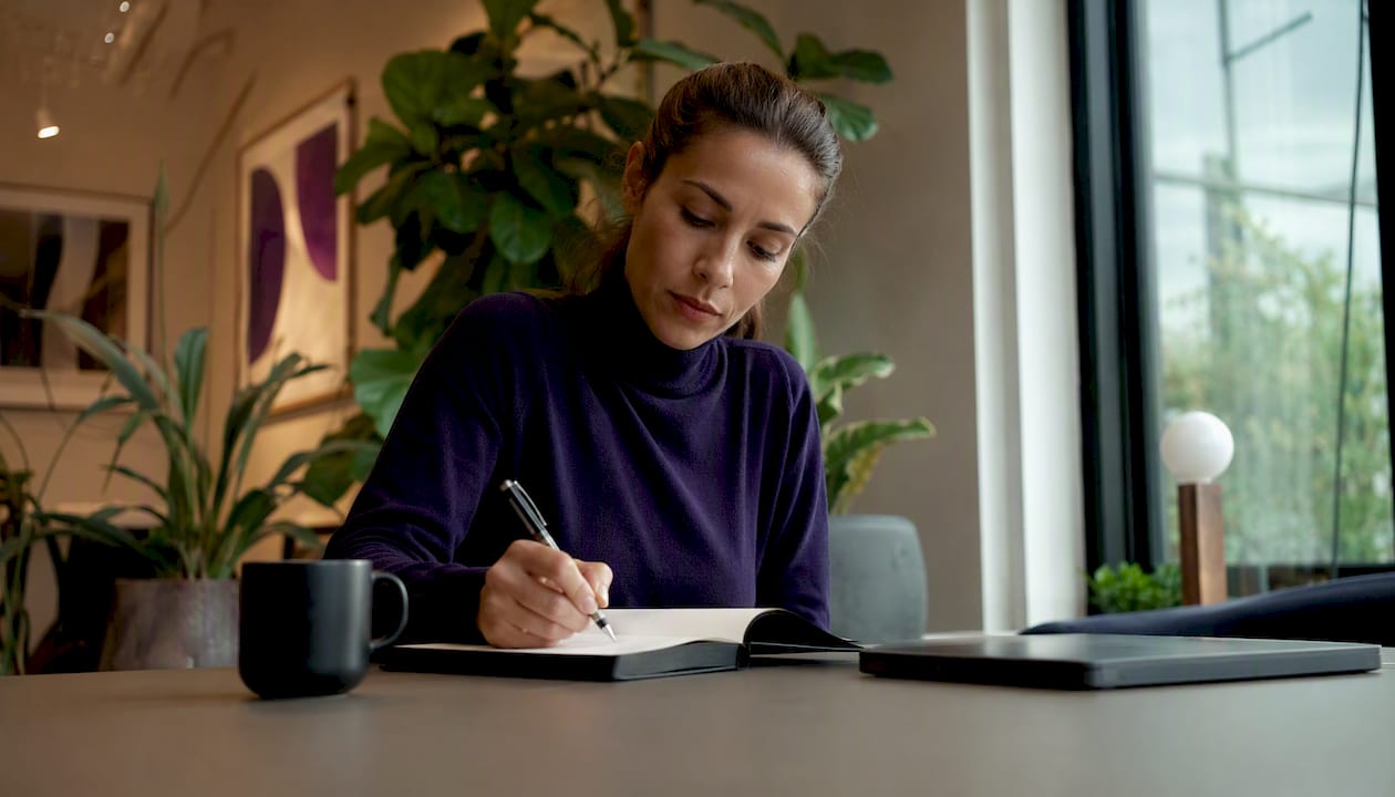 En kvinna sitter vid sitt skrivbord hemma och antecknar på ett papper.
