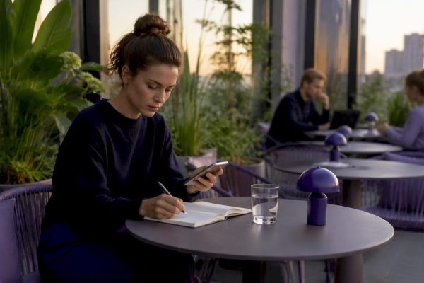 En kvinna sitter och jobbar i ett coworking-space med mobilen i handen och anteckningsblocket framför sig.