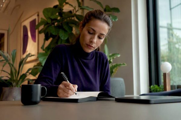 En kvinna sitter vid sitt skrivbord hemma och antecknar på ett papper.